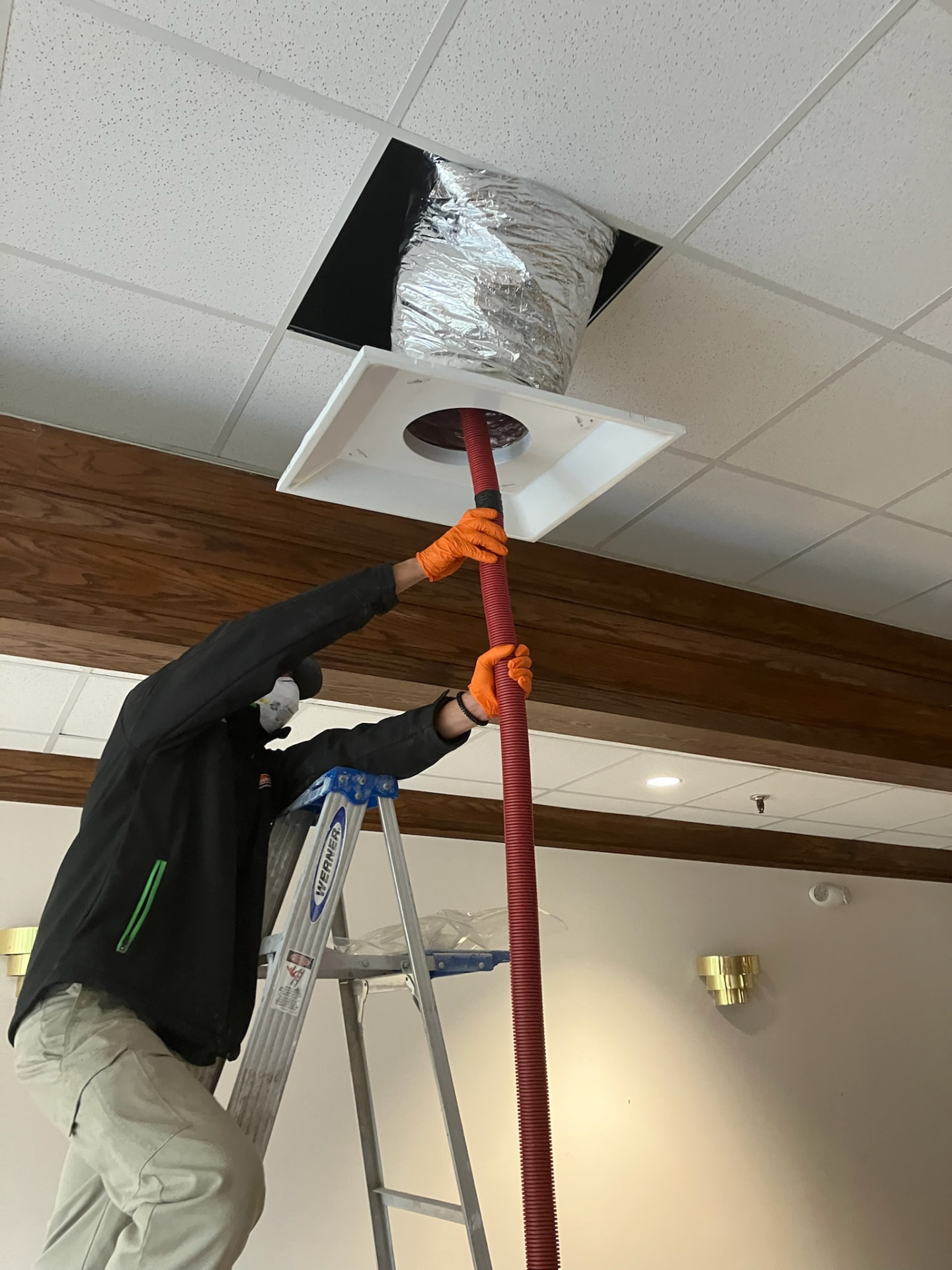 IEI technician on a ladder connecting a vacuum hose to an above-ceiling air duct opening during remediation work