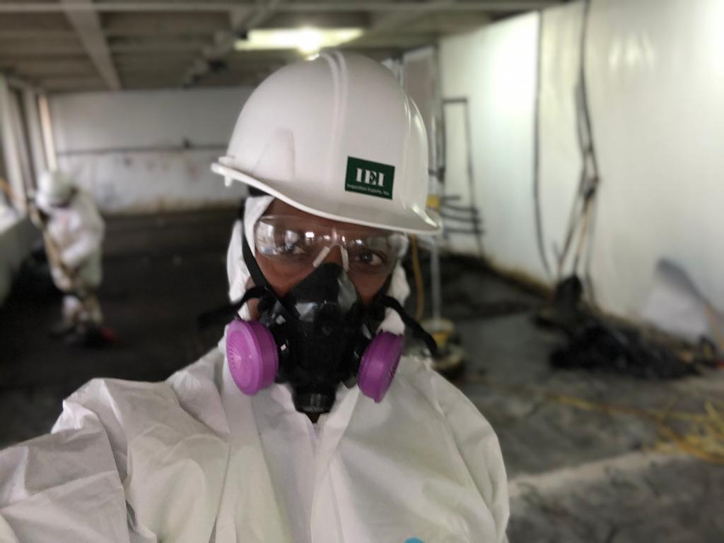 IEI worker in a respirator, protective suit, and IEI hard hat inside an abatement work area