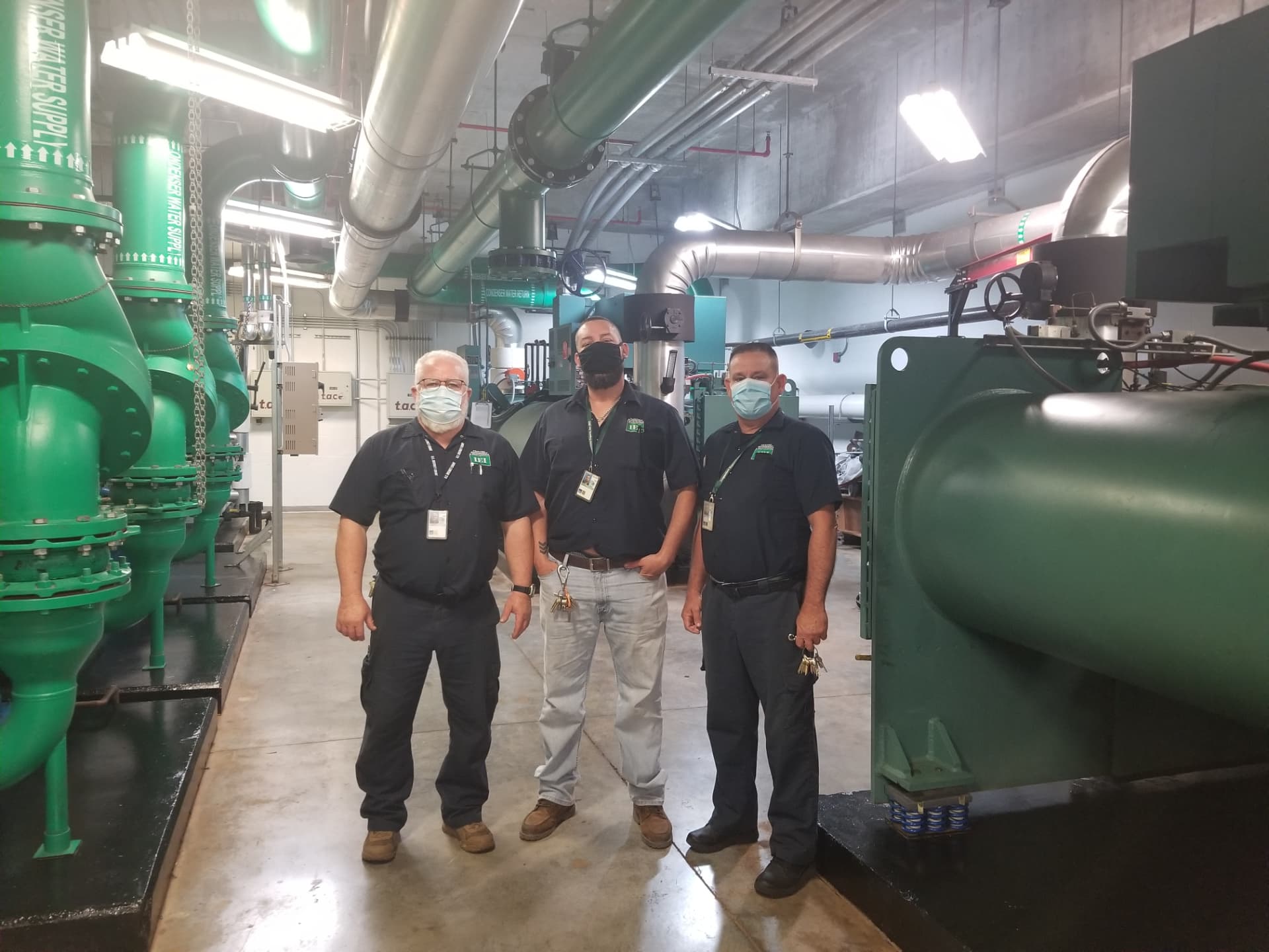 Three IEI team members standing in a central chiller plant with green condenser water piping